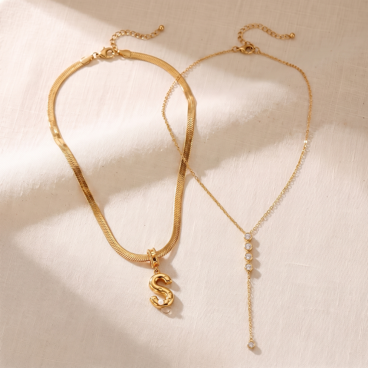 Il Look Alma: la collana con iniziale e perla e la collana a Y con zirconi appoggiate insieme su lino crema
