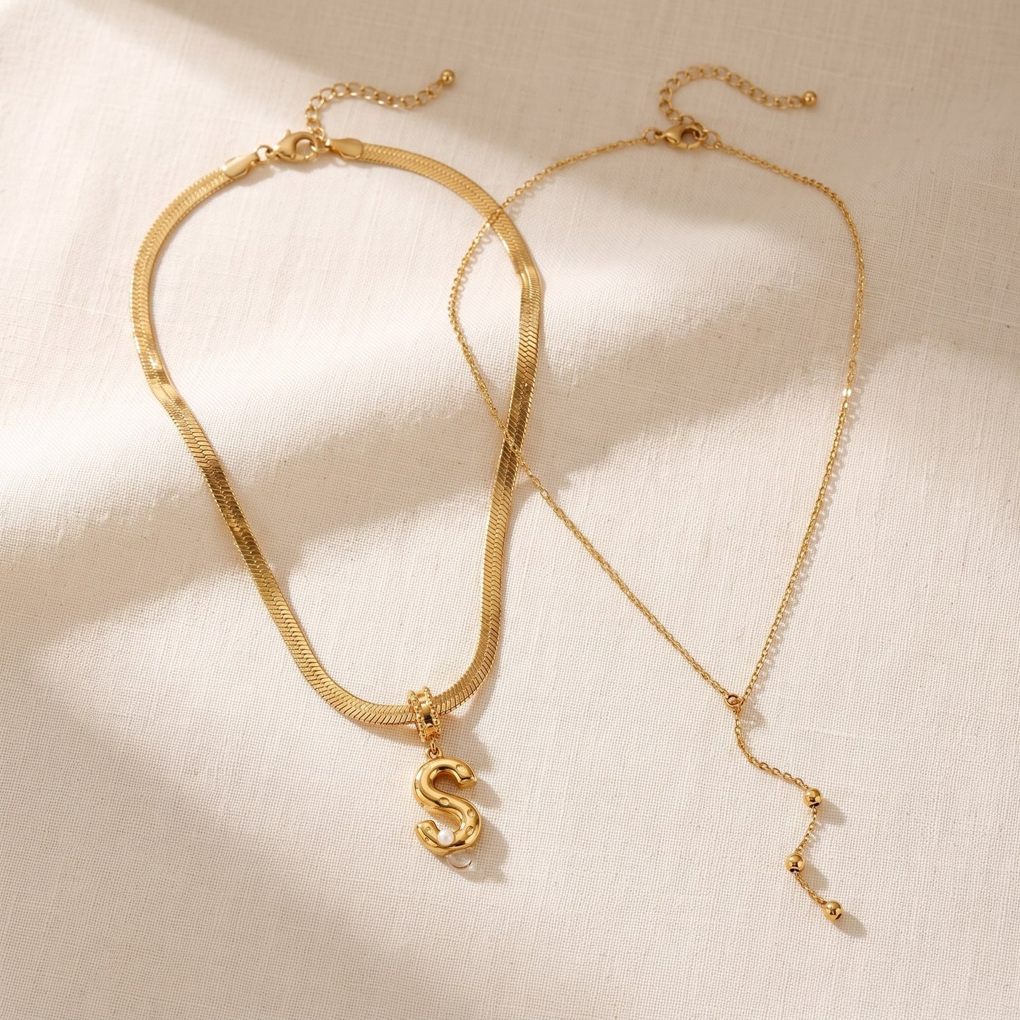 Il Look Alma: la collana con iniziale e perla e la collana a Y sottile appoggiate insieme su lino crema