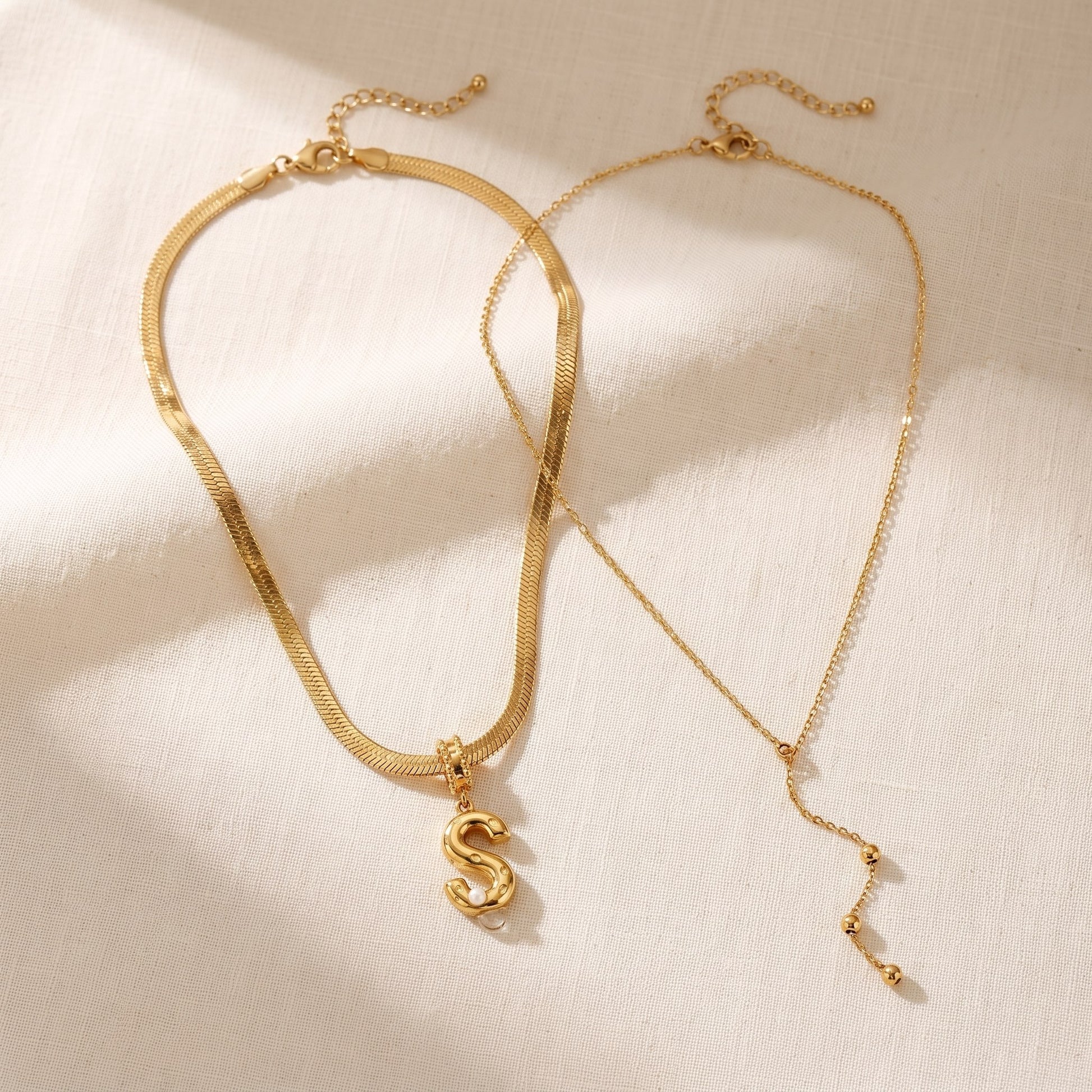 Il Look Alma: la collana con iniziale e perla e la collana a Y sottile appoggiate insieme su lino crema