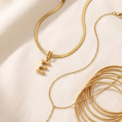 Il Look Completo: collana con iniziale, collana a Y e bracciali rigidi appoggiati insieme su lino crema